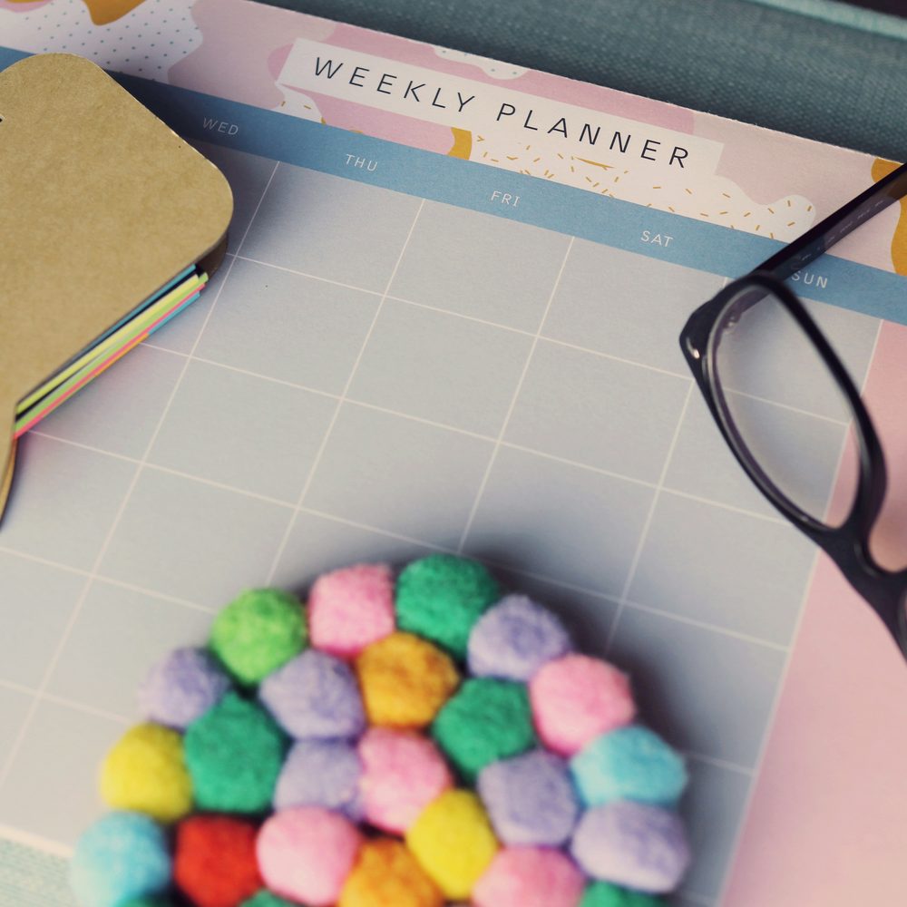 A weekly planner page on a desk beside a notebook, reading glasses and a cluster of coloured pom-poms