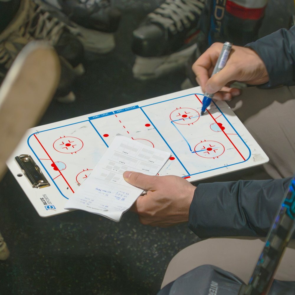 A coach's hands marking up a rink board and a handwritten line-up sheet, with players' skates behind