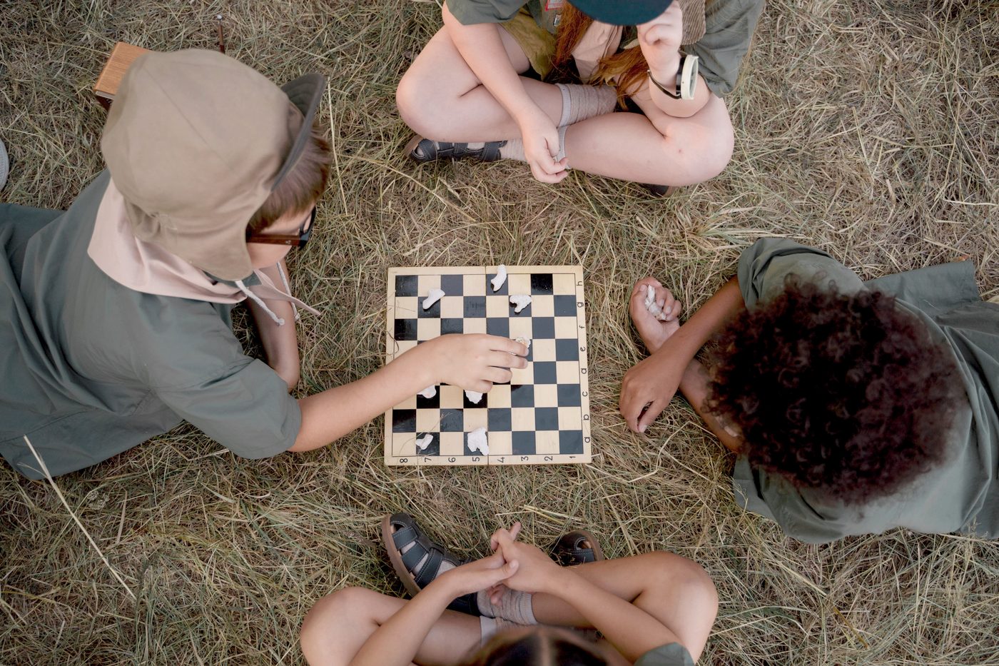Four children sitting cross-legged on dry grass around a wooden chessboard, seen from above